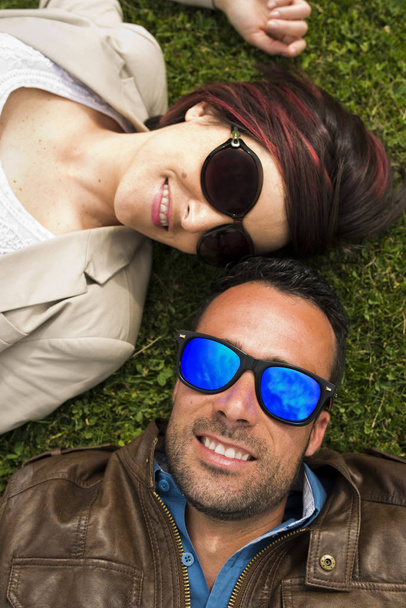 Aurinkolasit pari poseeraa kaupungin puistossa hymyillen nurmikolla
 - Valokuva, kuva