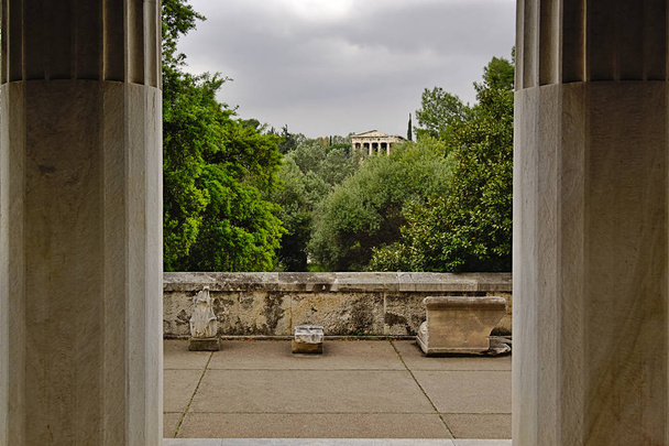 View of trees, temple of Hephaestus and cloudy sky between two columns in Stoa of Attalos in Athens, Greece. Impressive building detail in Ancient Agora archeological site. - Photo, Image