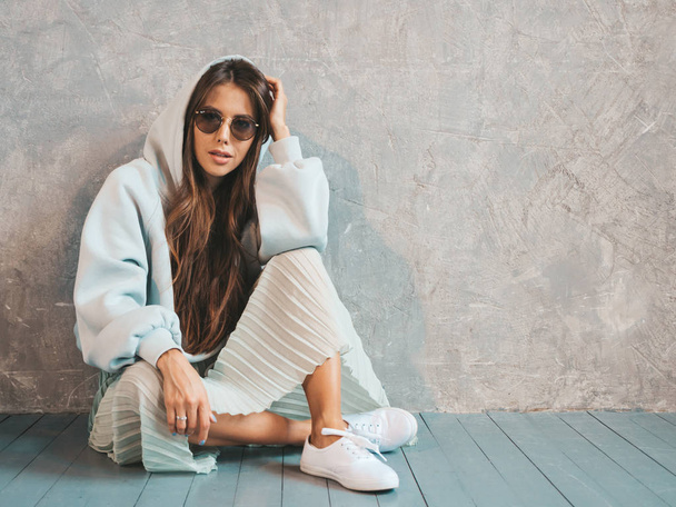 Young beautiful woman looking at camera. Trendy girl in casual summer hoodie and skirt clothes. Funny and positive female posing near gray wall in studio. In sunglasses sitting on the floor - Foto, imagen