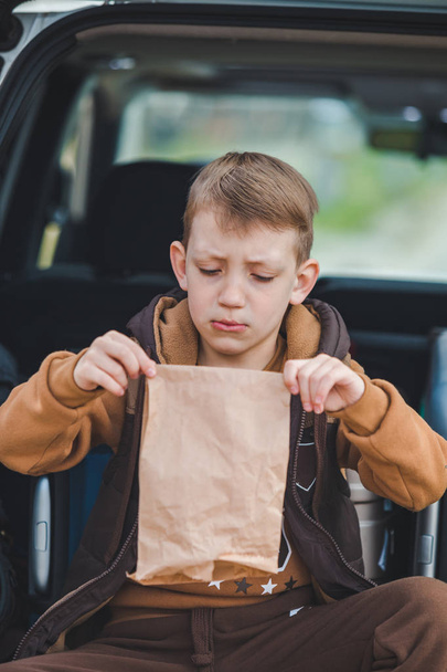 mały dzieciak patrząc w papierową torbę z cukierkami siedzi w bagażniku samochodu - Zdjęcie, obraz