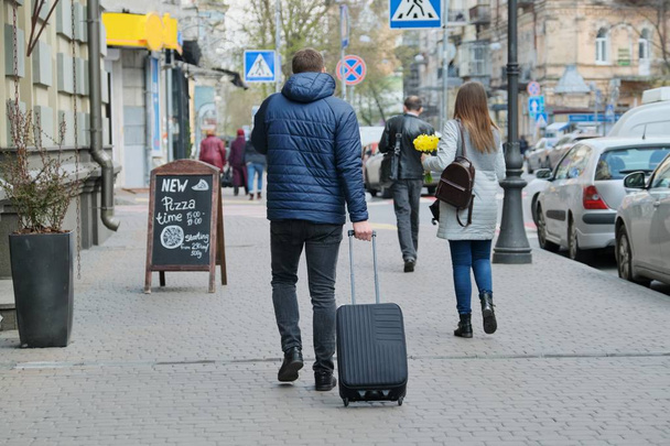 Человек в куртке с чемоданом, идущий по городской улице, вид сзади
 - Фото, изображение