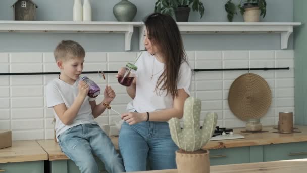 Young mom and son drinking juice with candy at kitchen. Happy mother spending time with son sitting on table in slow motion - Filmati, video