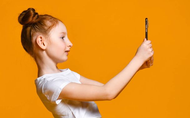 cheerful little red-haired girl taking selfie with mobile phone against a yellow wall. - Foto, imagen