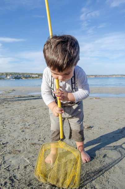 Schattige twee jaar oude jongen spelen met visnet op zonnige dag op het strand van de zee - Foto, afbeelding