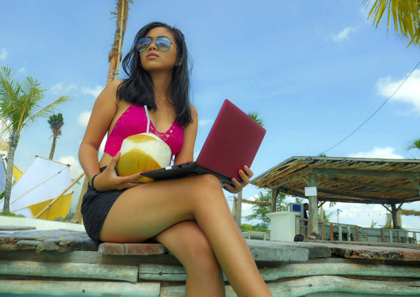 young beautiful and happy Asian Indonesian teenager woman in bikini at tropical resort swimming pool networking relaxed and chilled with laptop computer smiling - 写真・画像