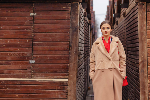 jovem elegante bonito menina em um casaco de pele passeando pela cidade perto de casas de madeira e paredes de pedra
 - Foto, Imagem