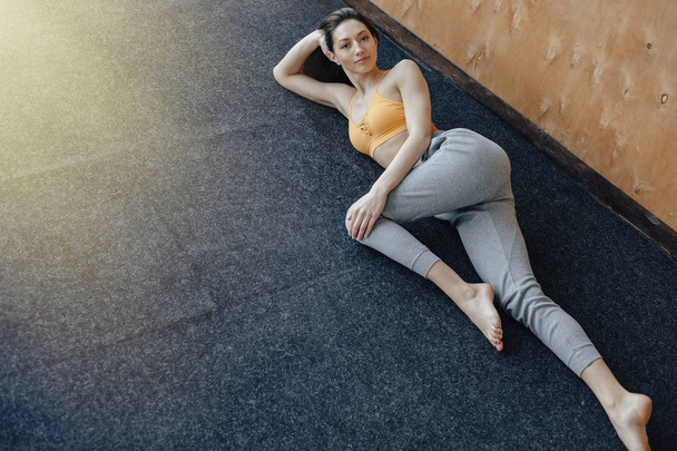Menina fitness atraente jovem deitado no chão perto da janela no fundo de uma parede de madeira, descansando em aulas de ioga
 - Foto, Imagem