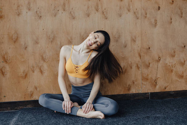 Jonge aantrekkelijke fitness meisje zittend op de vloer in de buurt van het raam op de achtergrond van een houten muur, rust op yogalessen - Foto, afbeelding