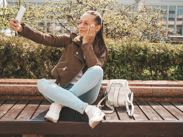 Retrato de bela menina morena sorridente no verão casaco hipster e jeans.Model tirar selfie no smartphone.Woman fazendo fotos em dia ensolarado quente na rua. Sentado no banco de óculos de sol
 - Foto, Imagem