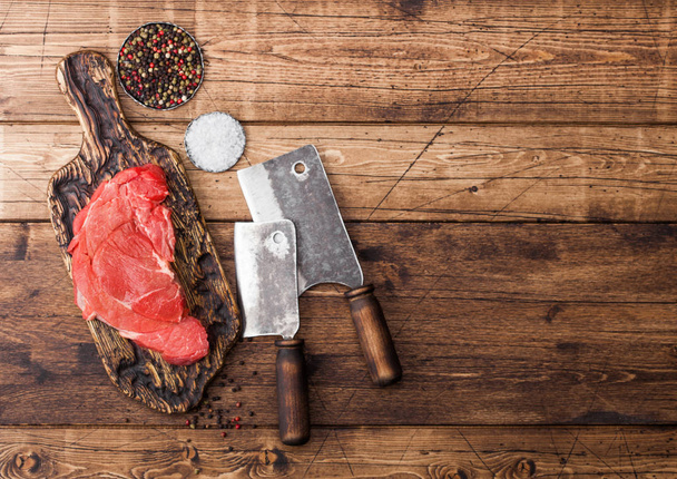Fresh raw organic slice of braising steak fillet on chopping board with meat hatchets on wooden background. Salt and Pepper - Photo, Image
