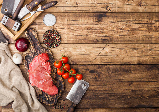 Fresh raw organic slice of braising steak fillet on chopping board with meat hatchet on wooden background. Red onion, tomatoes with salt and pepper. Space for text - Photo, image