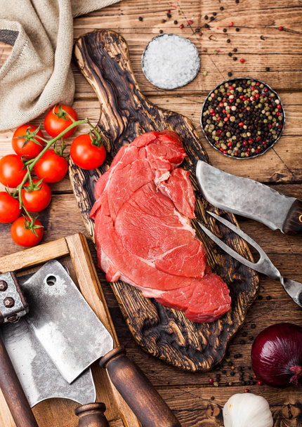 Fresh raw organic slice of braising steak fillet on chopping board with fork and knife on wooden background. Red onion, tomatoes with salt and pepper. - Foto, immagini