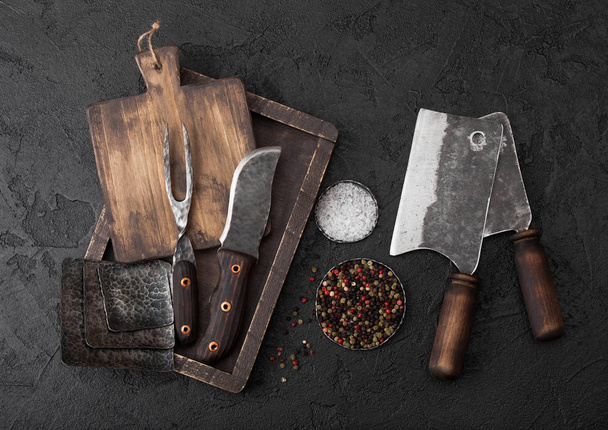 Vintage meat knife and fork and hatchets with vintage chopping board and black table background. Butcher utensils. Close up. Salt and pepper - Foto, Imagem