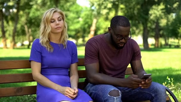 African american man playing cellphone on date in park, ignoring sad girlfriend - Valokuva, kuva