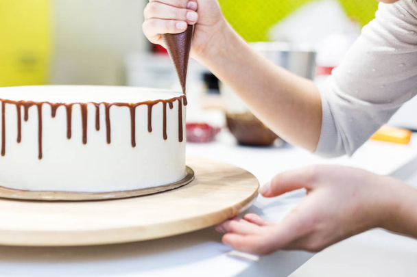 A confectioner squeezes liquid chocolate from a pastry bag onto a white cream biscuit cake on a wooden stand. The concept of homemade pastry, cooking cakes. - Foto, Imagem