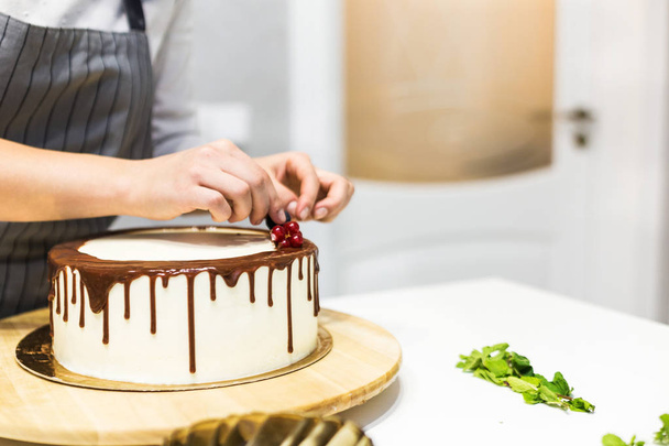 Confectioner decorates with berries a biscuit cake with white cream and chocolate. Cake stands on a wooden stand on a white table. The concept of homemade pastry, cooking cakes. - Fotó, kép