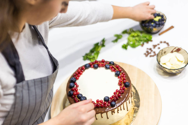 Confectioner decorates with berries a biscuit cake with white cream and chocolate. Cake stands on a wooden stand on a white table. The concept of homemade pastry, cooking cakes. - Photo, image
