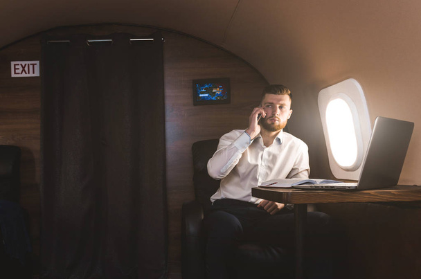 Young attractive and successful businessman talking on the phone and working while sitting in the chair of his private business plane - Zdjęcie, obraz