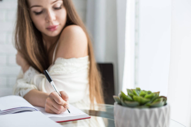 Piękna młoda dziewczyna w koszuli nocnej siedzi rano na szklanym stole i pisze jej myśli w Notatniku. - Zdjęcie, obraz