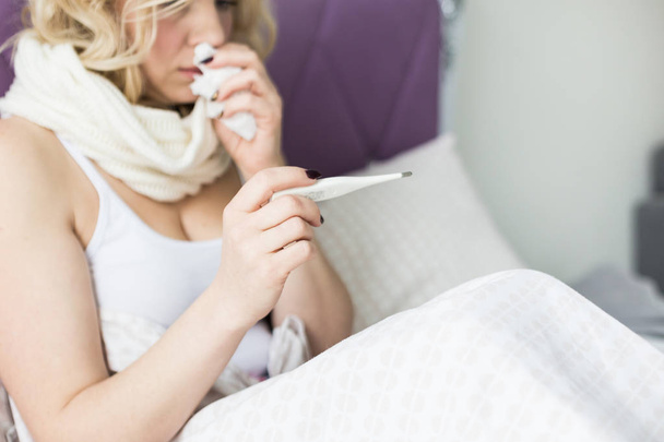 Beautiful blond woman sitting in bed with a thermometer and a scarf wrapped around the neck and a handkerchief. illness and health at home - Foto, imagen
