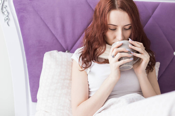 An attractive young brunette woman sits in her bed with a mug and pills. Colds and home treatment - Foto, imagen