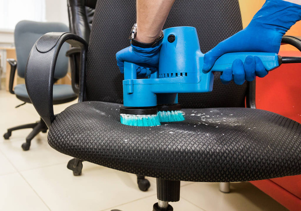 Young man in workwear and rubber gloves cleans the office chair with professional equipment. - Zdjęcie, obraz