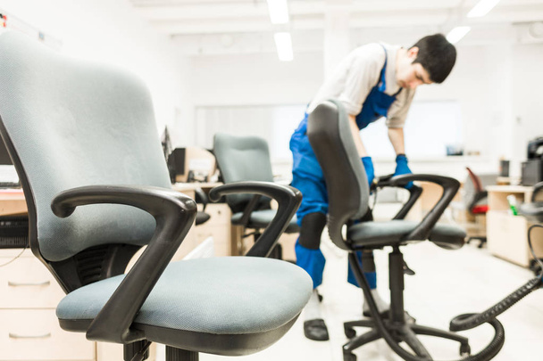 Young man in workwear and rubber gloves cleans the office chair with professional equipment. - Foto, Imagen