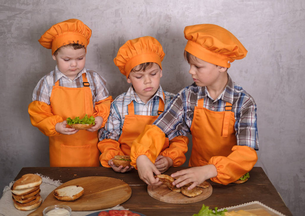 three cute boys in costumes cooks engaged in cooking homemade burgers. three brothers preparing family dinner - Photo, Image