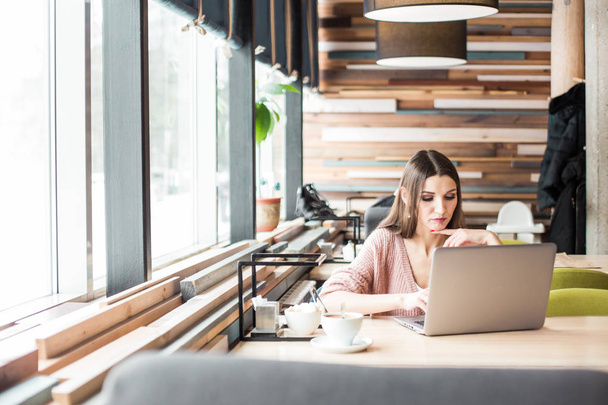 Aantrekkelijke jonge vrouw zit aan een tafel in een cafe met een kopje koffie en geniet van een laptop - Foto, afbeelding