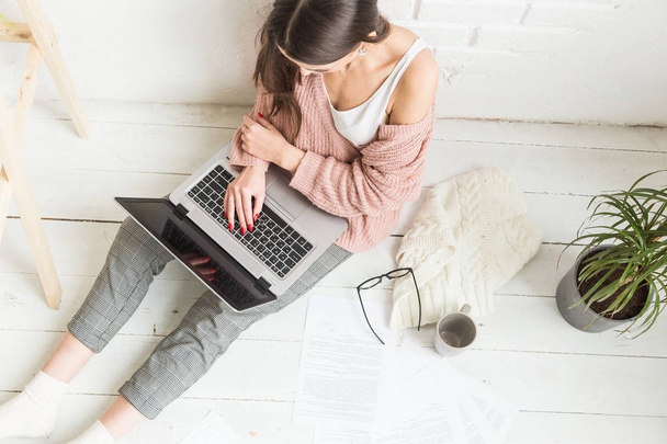 A young happy woman sits on the floor in a bright apartment or office interior and works at a laptop, freelancer girl at work, the concept of distance learning students, paperwork and online learning - Фото, изображение
