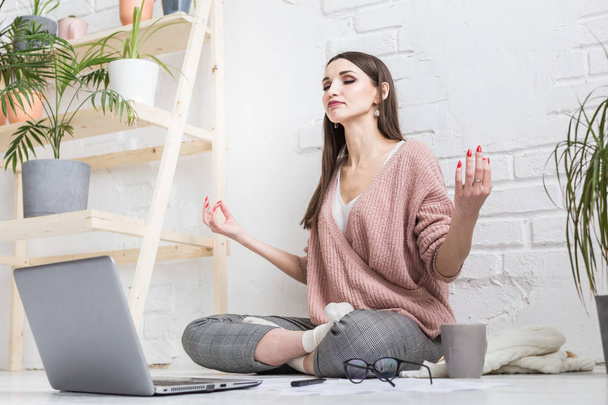 Young happy woman sits on the floor in a yoga pose in a bright apartment and works behind a laptop, a freelancer girl meditates while working. control stress - Photo, image