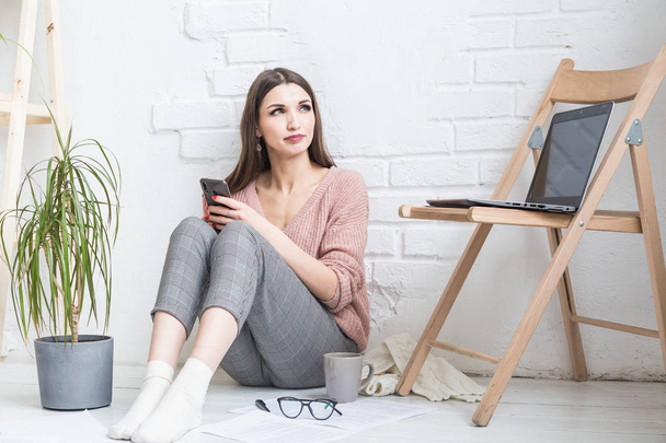 A happy woman in homemade casual clothes sits in a bright interior and uses a telephone, the girl is resting while working at a laptop and surfing the Internet. Cozy freelance work space, - Foto, afbeelding