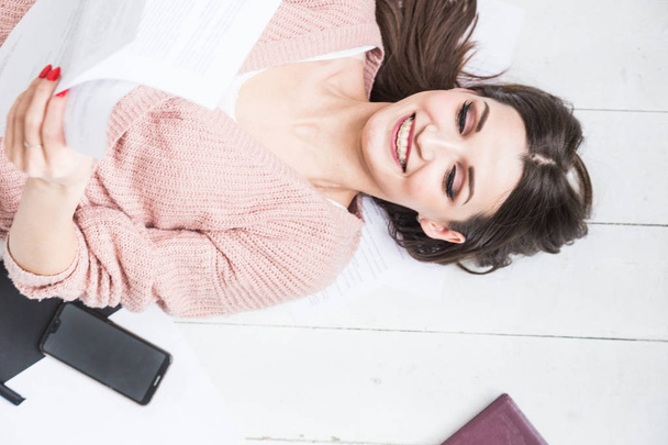 A beautiful woman lies on the floor among papers and documents, the girl freelancer smiles and relaxes during a break from work - Foto, Imagem