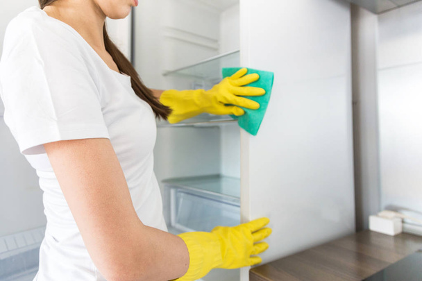 Nuori nainen siivousfirmasta siivoaa kotona. Mies pesee keittiön pesee jääkaapin keltaisilla hanskoilla siivoustarvikkeilla. - Valokuva, kuva