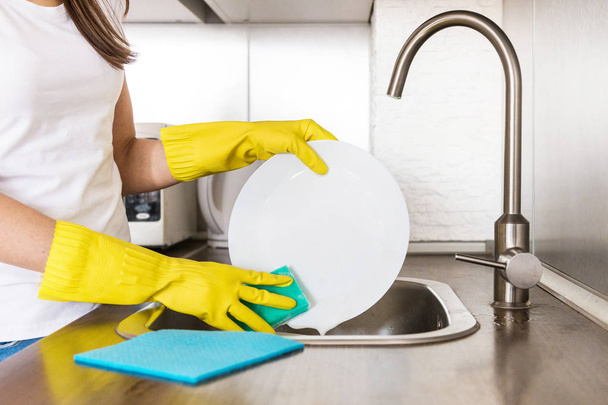 Een jonge vrouw met gele handschoenen wast de vaat met een spons in de gootsteen. Professionele schoonmaakdienst. - Foto, afbeelding