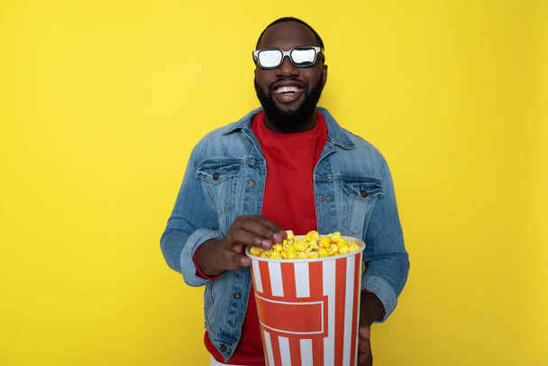 Waist up of smiling African guy holding bucket with popcorn indoors - Φωτογραφία, εικόνα