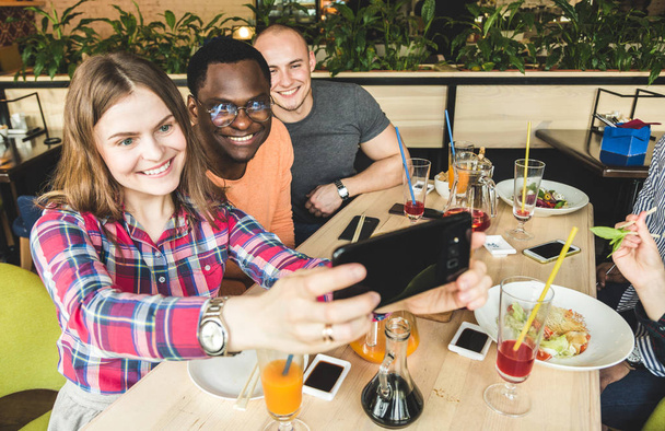 Ομάδα νεαρών χαρούμενων φίλων κάθονται σε ένα καφέ, τρώγοντας, πίνοντας ποτά. Οι φίλοι βγάζουν selfies και βγάζουν φωτογραφίες. - Φωτογραφία, εικόνα