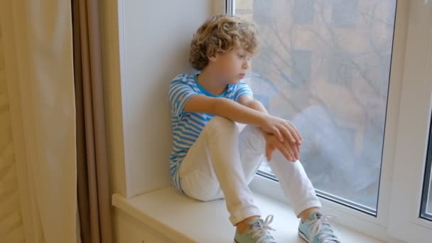 窓枠の上に座っているカーリーブロンドの少年が窓の外を見る。6歳の子供は寝室に一人で座って悲しい - 映像、動画
