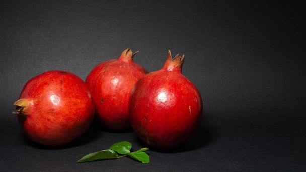 Bright juicy pomegranate fruit, black background, in the photo three fruits located in the lower left corner, with green foliage, close-up - 写真・画像