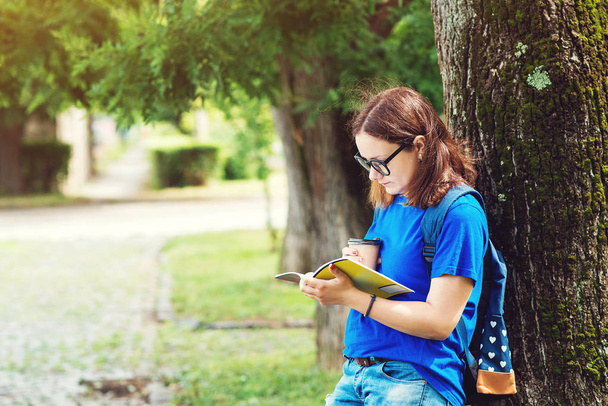 Красивая студентка в очках, читает блокнот, пьет кофе в кампус-парке
.  - Фото, изображение