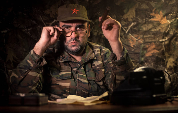 El malvado dictador sentado en la mesa. General comunista enojado sentado en el cuartel general o comandante cubano en cuarto oscuro. Estudio decoración
 - Foto, Imagen