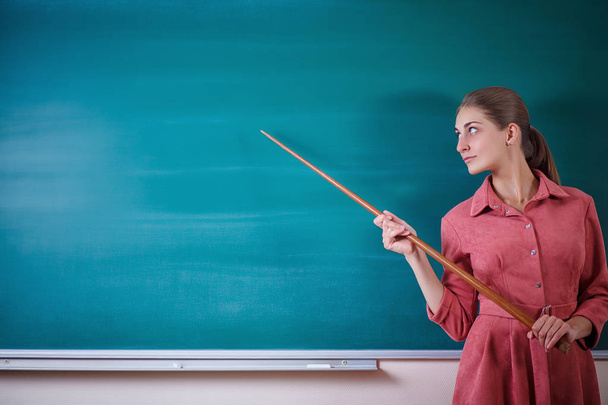 若い女性教師はポインター付きの黒板に立っている。学校の時刻表やその他の情報のテンプレート。美しいタッチャーは、教育委員会の空のコピースペースに表示されます。教育の概念. - 写真・画像
