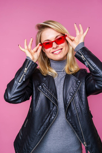 Photo of happy blonde in pink glasses and leather jacket with hands near face - Fotografie, Obrázek