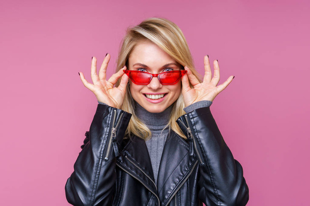 Image of happy blonde in pink glasses and leather jacket with hands near face - Photo, Image