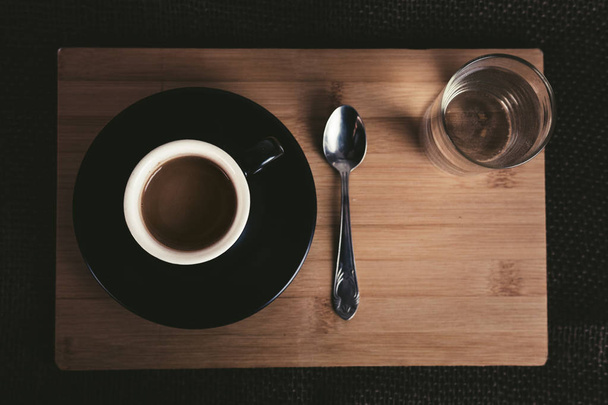 Photo of illuminated small black coffee cup on wooden table with tea spoon and glass full of clear water on dark background. Espresso on the wooden board with natural light, from above - top view. - Foto, Imagem