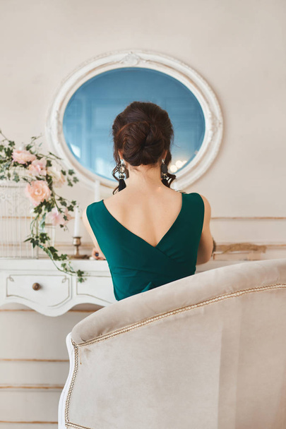 Beautiful brunette girl in dress sits back to camera and looks in mirror - Фото, изображение
