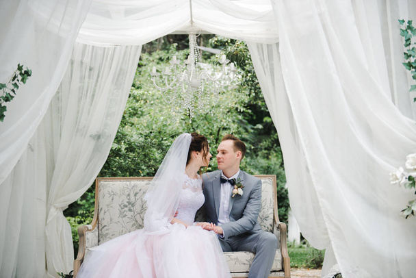 Hochzeit. Braut und Bräutigam sitzen auf einer schönen Couch in einem Pavillon im Garten. die Braut im rosa Kleid, der Bräutigam im grauen Anzug. Weißes Zelt. Braut und Bräutigam spazieren im Wald. - Foto, Bild