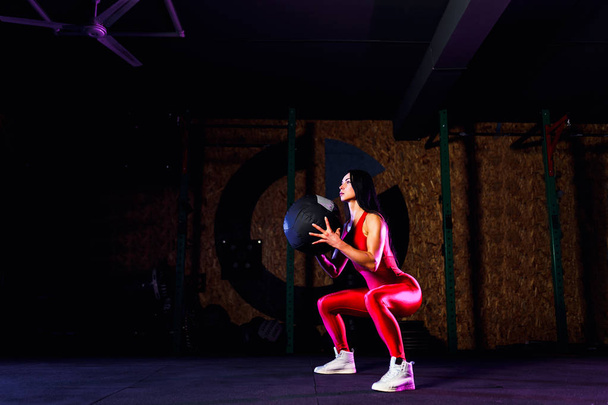 Sportswoman doing squat exercises with fitness ball. Female exercising and stretching with medicine ball at gym - 写真・画像