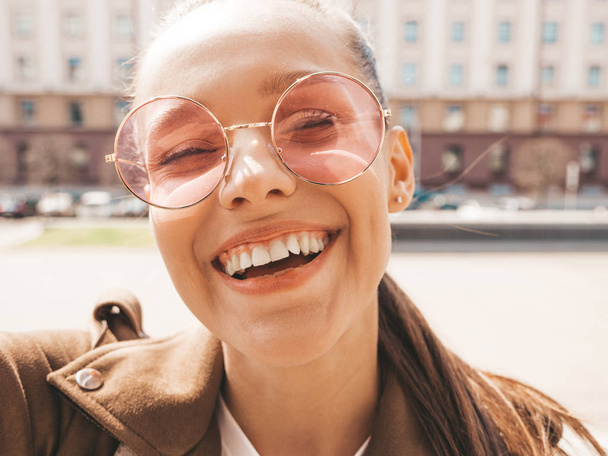 Closeup portrait of beautiful smiling brunette girl in summer hipster jacket. Model taking selfie on smartphone.Woman making photos in warm sunny day in the street in sunglasses - Photo, Image