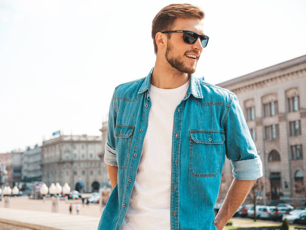 Portrait of handsome smiling stylish hipster lumbersexual businessman model. Man dressed in jeans jacket clothes. Fashion male posing on the street background in sunglasses - Zdjęcie, obraz